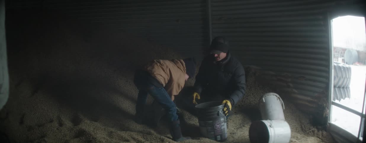 A father and son work together in slow motion, scooping dusty grain into a bucket inside a large metal silo