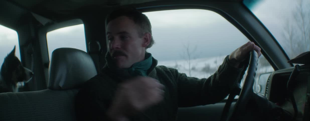 A mustachioed man in a cowboy hat gets into his truck, greets his dog, and begins a peaceful drive through a snowy, rural landscape