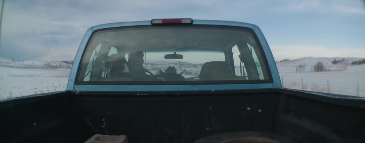 A rancher and his trusty dog drive their pickup truck through a snowy, desolate landscape