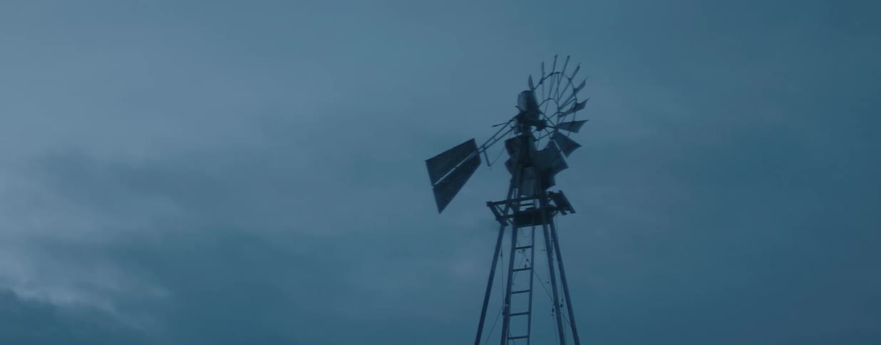 A silhouetted windmill slowly turns against a dark, stormy sky at dusk