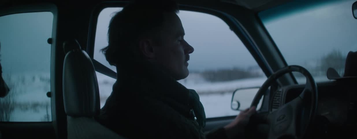 Side profile of a man with a mustache driving his truck through the snowy countryside at dusk