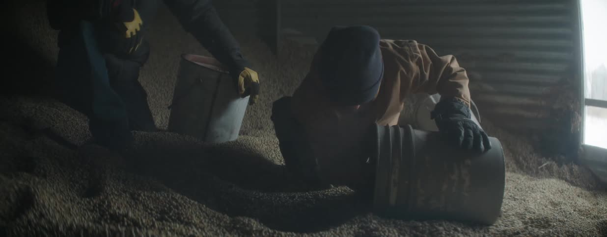 A father and son work together in slow motion, scooping grain into buckets inside a dimly lit silo