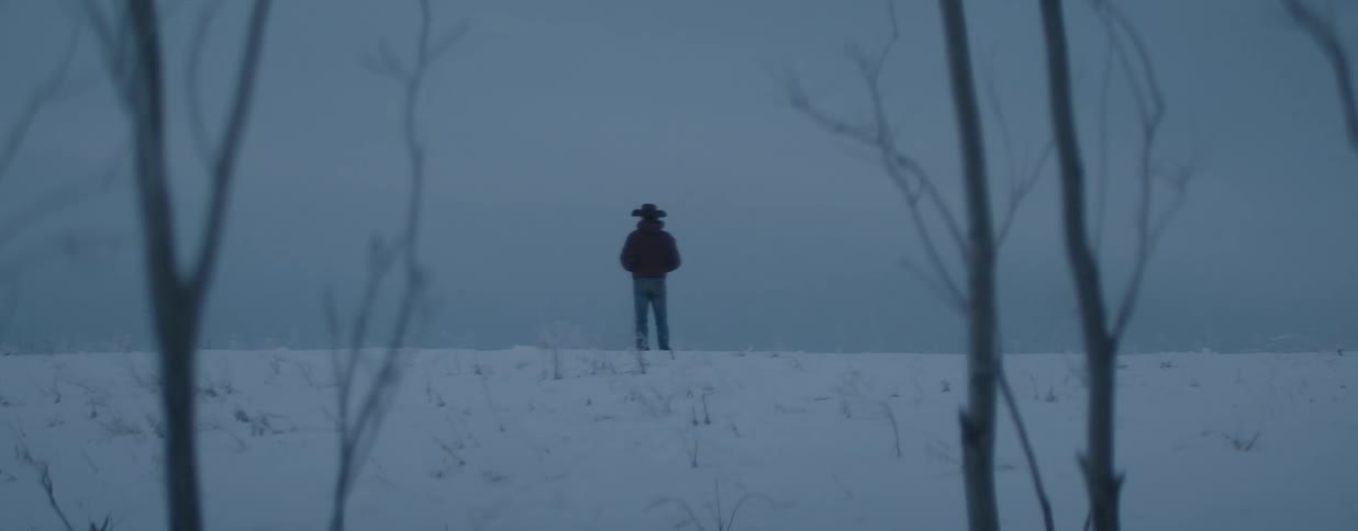 A solitary cowboy stands still in a vast, snow-covered field on a cold and gloomy winter day