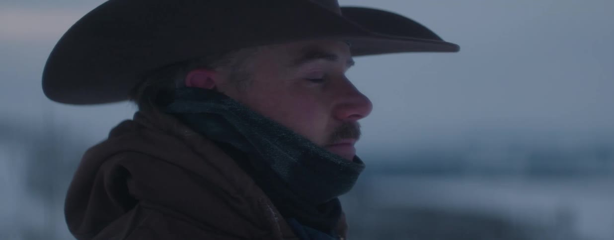 A thoughtful cowboy with frost on his scarf gazes into the distance on a bleak, cold winter day