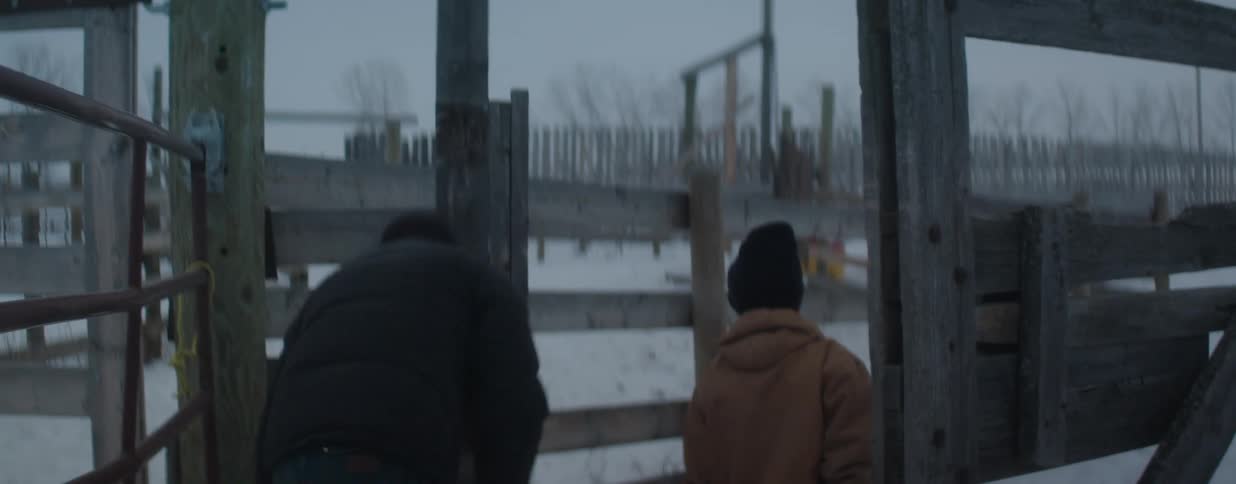 A rancher and his son walk together through a wooden gate into a snowy corral on a cold winter day
