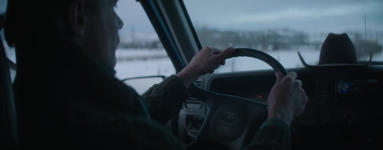 An over-the-shoulder view of a man driving a truck through a snowy, rural landscape on an overcast day