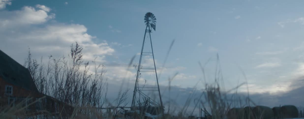 A classic farm windmill stands silhouetted against a cloudy twilight sky as the camera shifts focus to the dry winter grass in the foreground