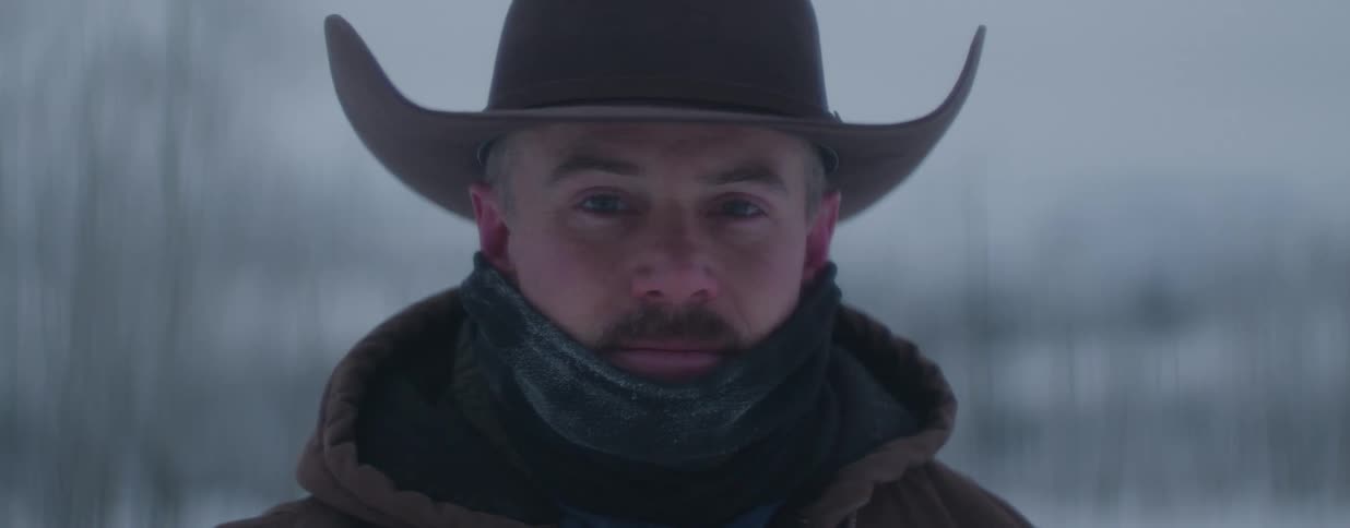 A stoic cowboy wearing a hat and scarf looks directly into the camera on a cold, snowy winter day