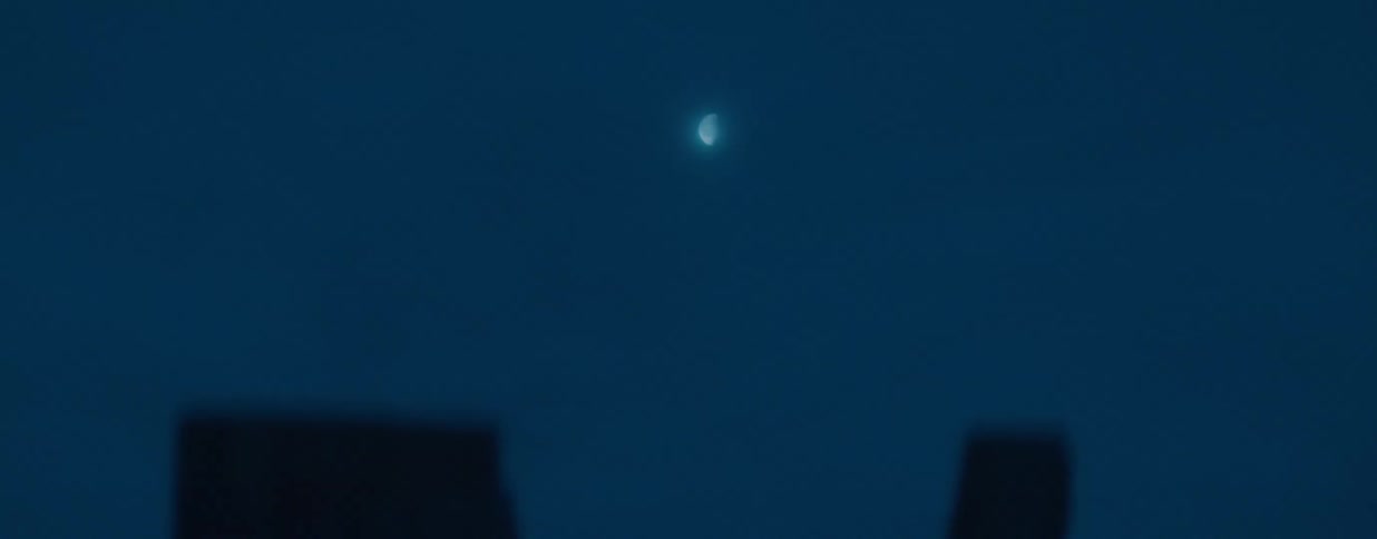 A crescent moon shines in the dark blue sky above the silhouette of buildings