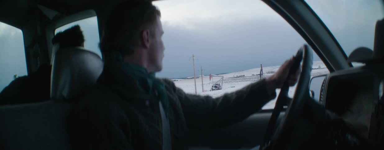 Interior shot of a man driving his car past a snowy, open field with oil pumpjacks in the distance