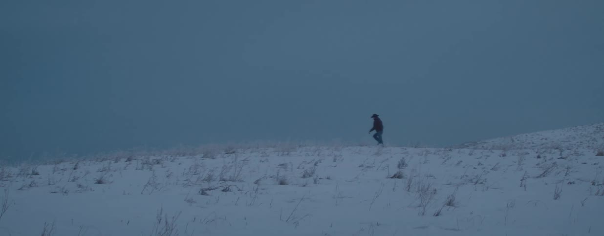 A lone cowboy walks across a desolate, snow-covered landscape during a cold and foggy winter dusk