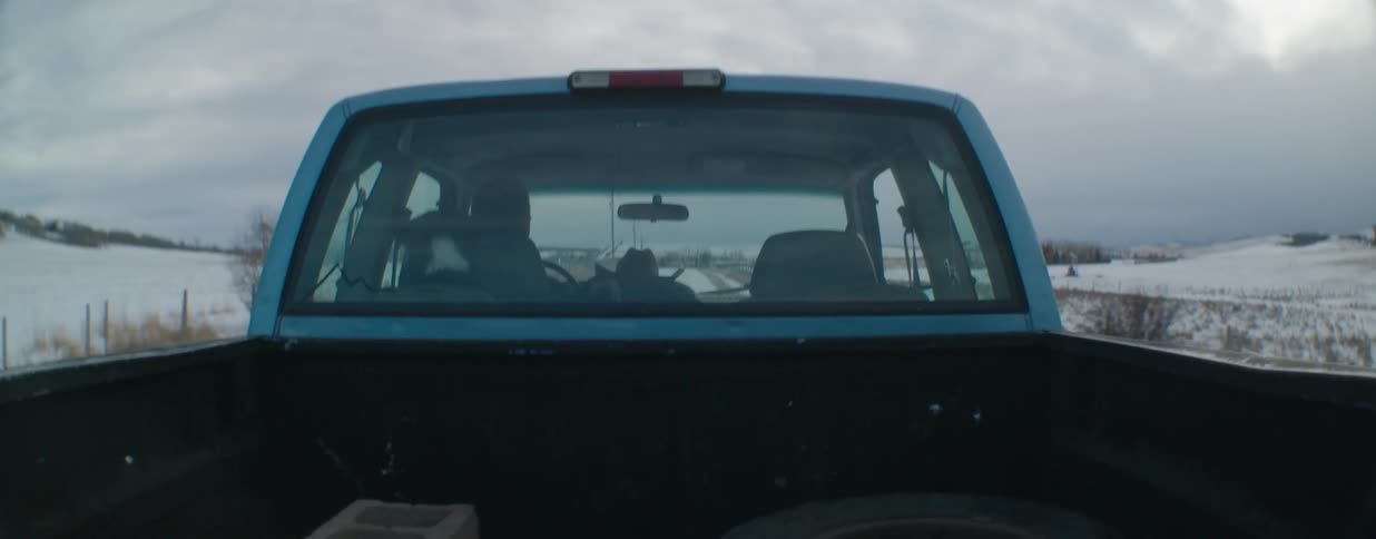 A cowboy and his dog drive their pickup truck down a snowy country road past oil fields on an overcast day