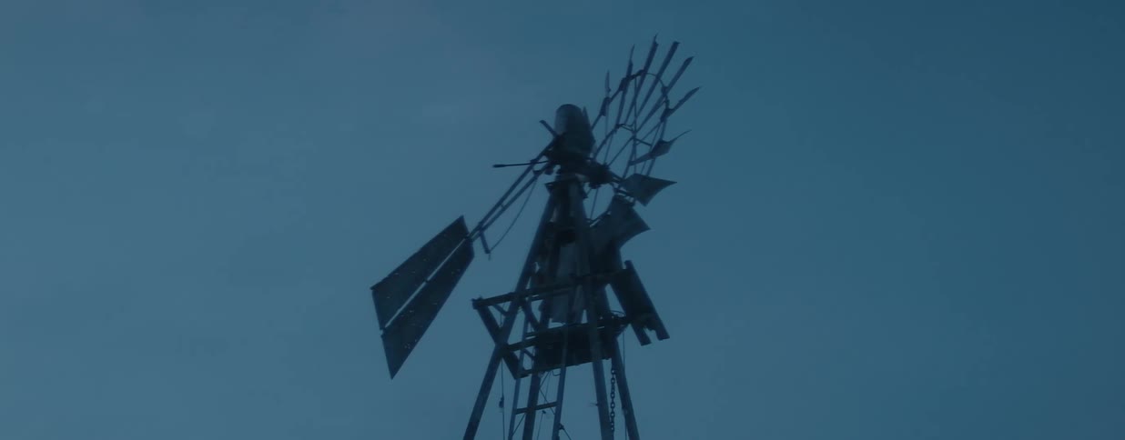 An old farm windmill stands silhouetted against a dark sky while snow falls gently at dusk