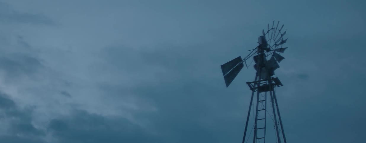A weathered farm windmill is motionless against a dark, overcast sky at twilight