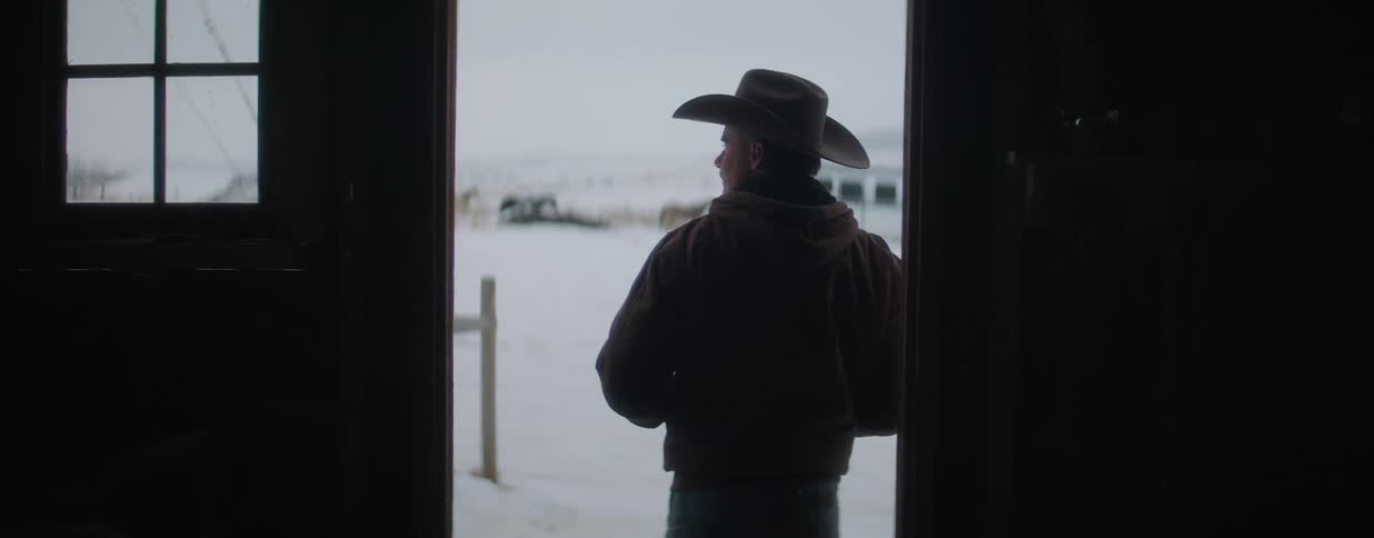 A cowboy stands silhouetted in a dark doorway, looking out at a snowy ranch landscape