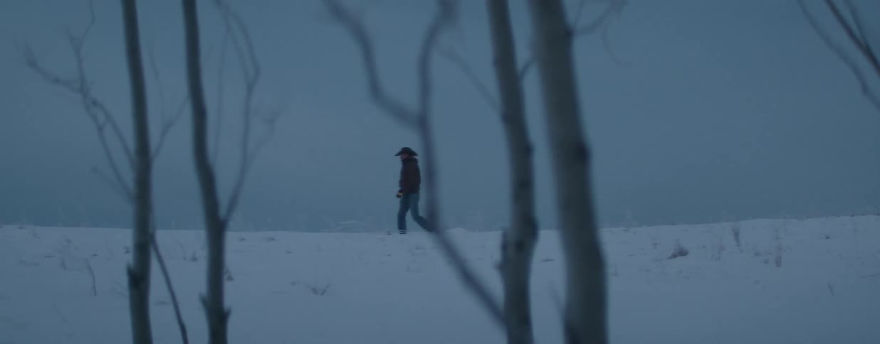 A man wearing a cowboy hat walks slowly through a vast, snowy landscape on a bleak and overcast winter day
