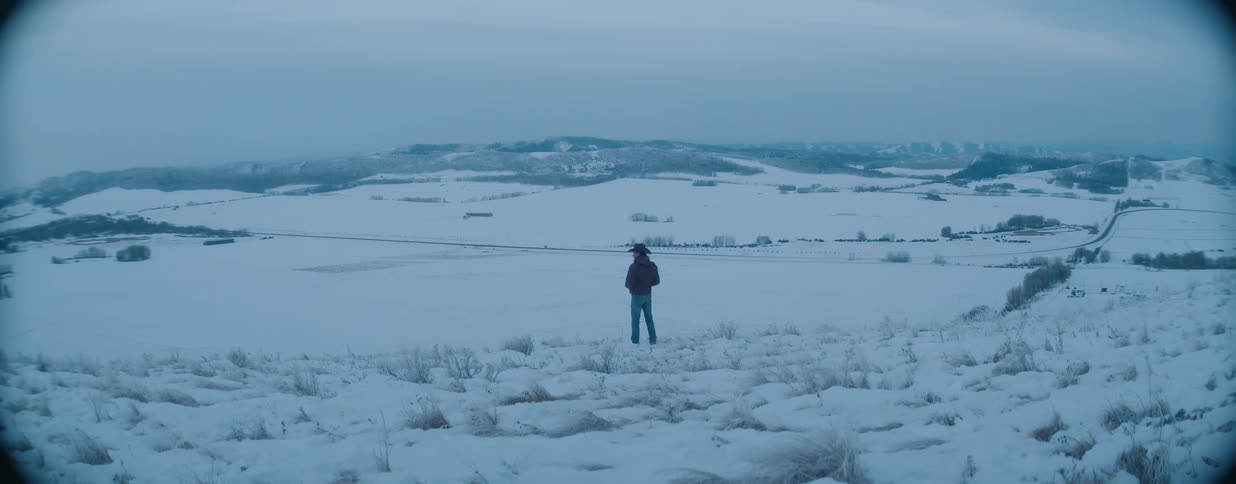 A lone cowboy in a winter coat walks to the edge of a snowy hill and looks out over the vast, frozen valley below
