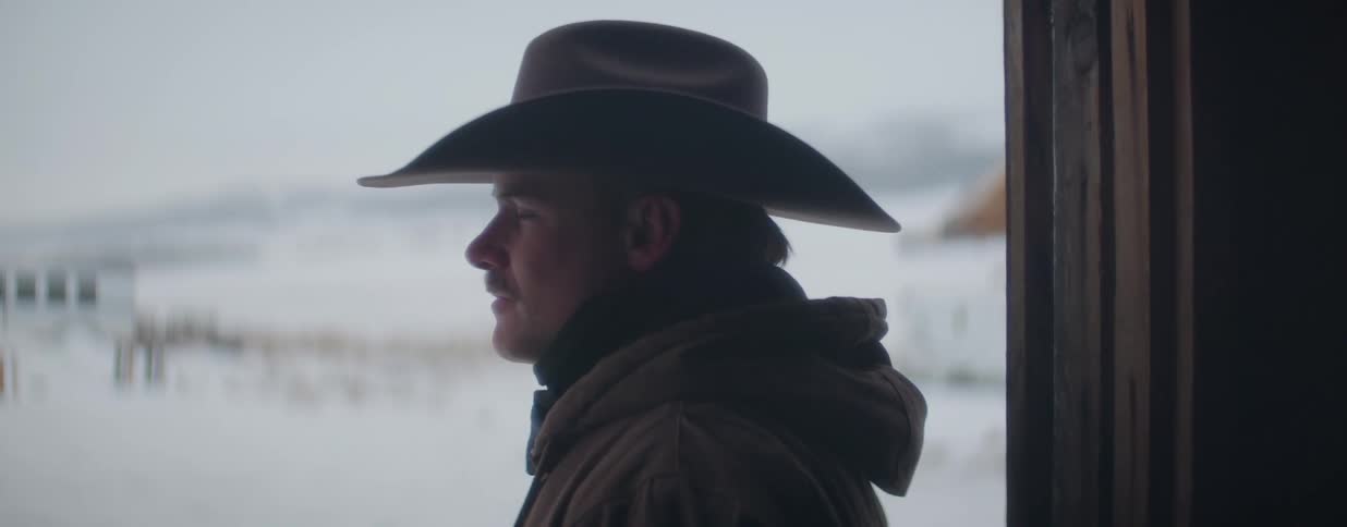 A thoughtful cowboy wearing a hat and jacket looks out over the snowy landscape from a doorway