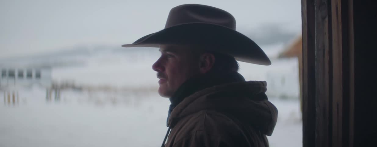 Close up profile of a handsome cowboy in a hat and jacket looking out over a snowy landscape on a cold winter day