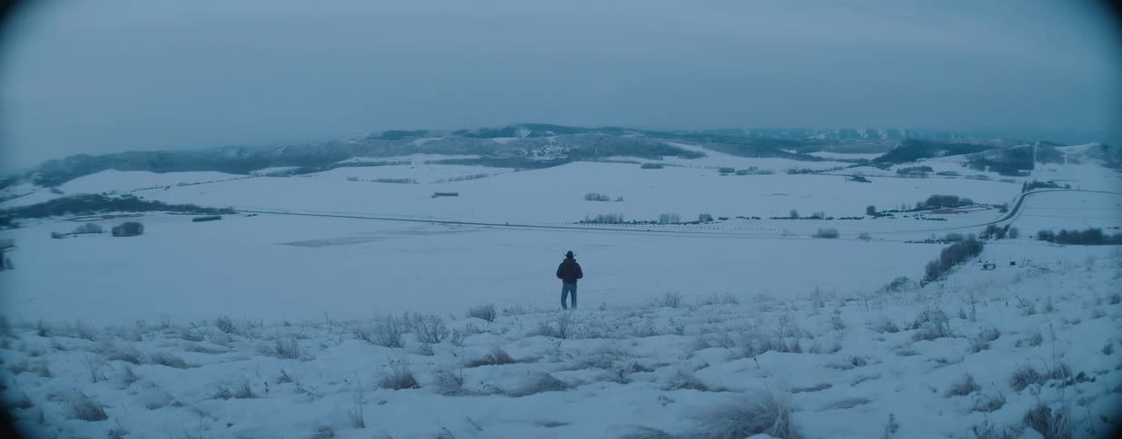 A man in a cowboy hat walks down a snowy hill, overlooking a vast, desolate valley on a cold winter day