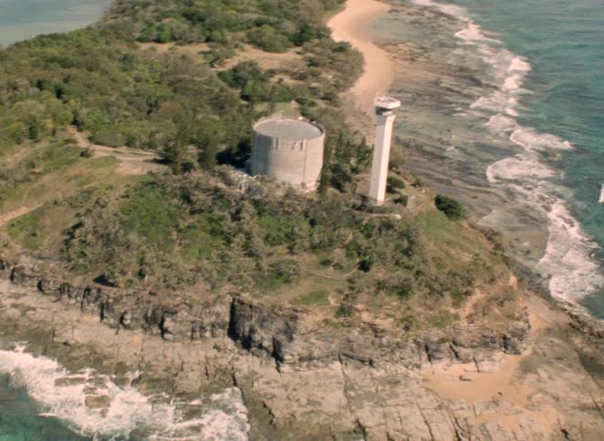 Vintage aerial footage reveals a lighthouse and a cylindrical building on a lush, rocky peninsula surrounded by the ocean