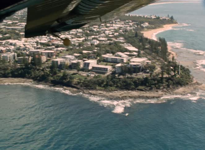A vintage aerial view from an airplane reveals a scenic coastal town with a rocky shoreline and a sandy beach