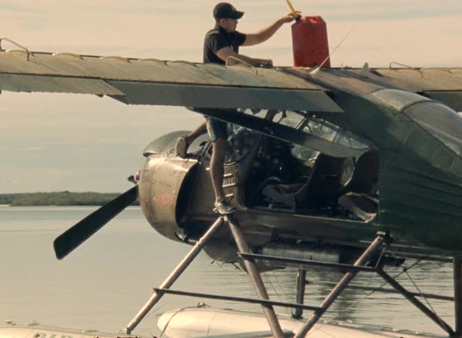 A man climbs onto the wing of a seaplane floating on the water to refuel it from a gas can