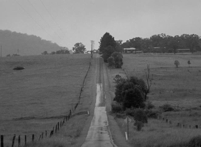 A vintage black and white film captures a long, wet country road stretching through rural fields on an overcast day