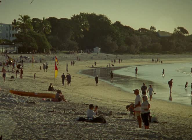 A nostalgic film-style shot shows a bustling beach scene at sunset with people walking and relaxing by the shoreline