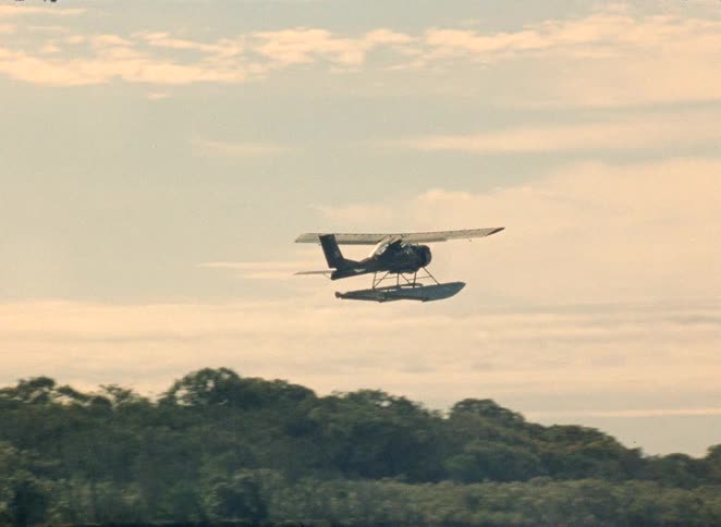 A vintage seaplane accelerates across the water, lifts off, and flies into the hazy sky