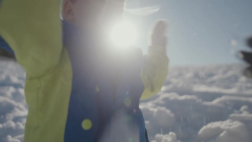 A toddler in a colorful snowsuit crumbles a snowball in slow motion while sitting in the snow on a bright sunny day