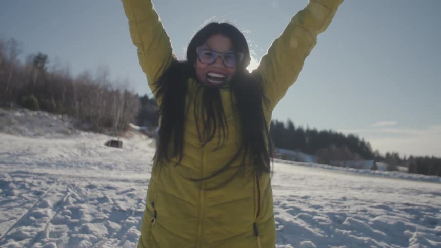 Slow motion shot of a joyous woman in a yellow winter coat throwing snow into the bright sunlight