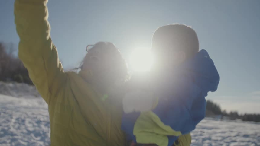 A joyful mother holds her toddler and playfully tosses snow into the air, watching it sparkle in the bright winter sun in slow motion