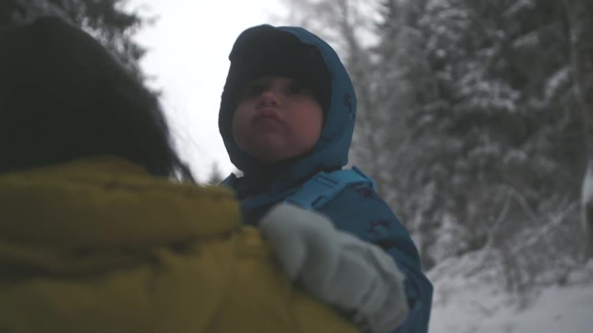 Over-the-shoulder view of a parent carrying their toddler who curiously observes the wintery forest scenery in slow motion