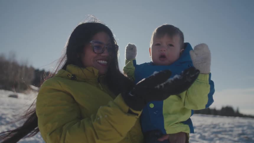 A smiling mother holds her baby son as she playfully blows snow into his face in slow motion on a sunny winter day
