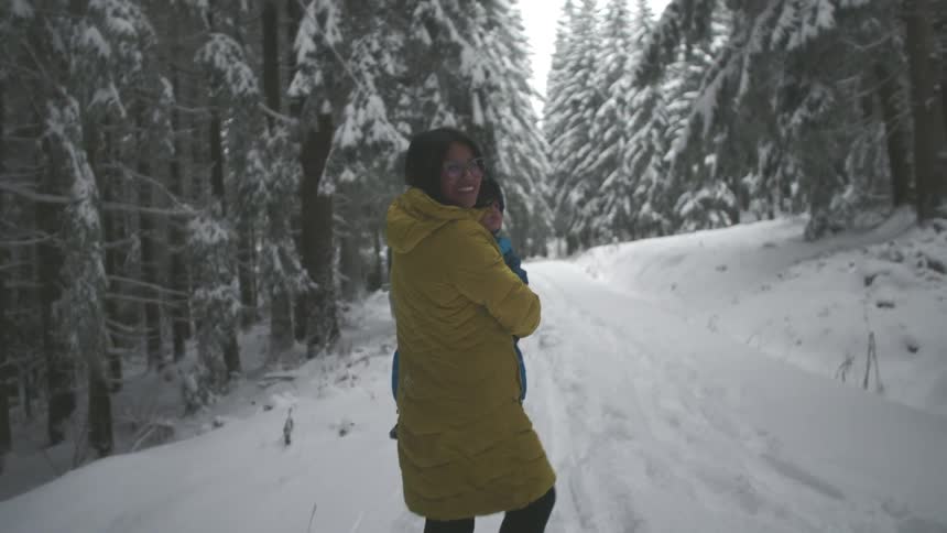 A smiling mother holds her young child in her arms while standing on a path in a beautiful, snow-covered forest