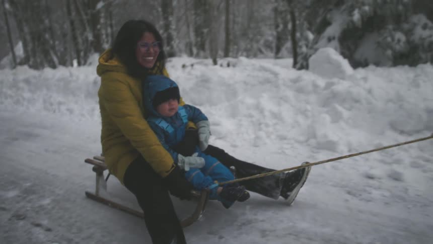 A joyful mother holds her toddler as they are pulled on a sled through a winter wonderland in slow motion