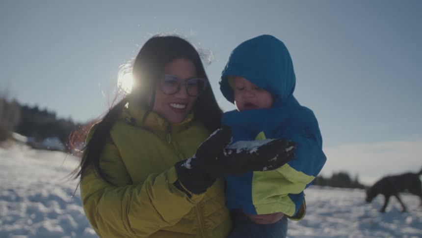 A smiling mother holds her bundled-up toddler as he touches snow for the first time on a bright, sunny winter day