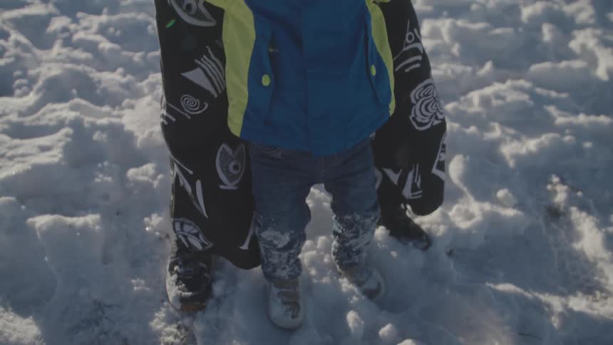 A toddler kicks their feet in the bright white snow in slow motion while being held by their parent