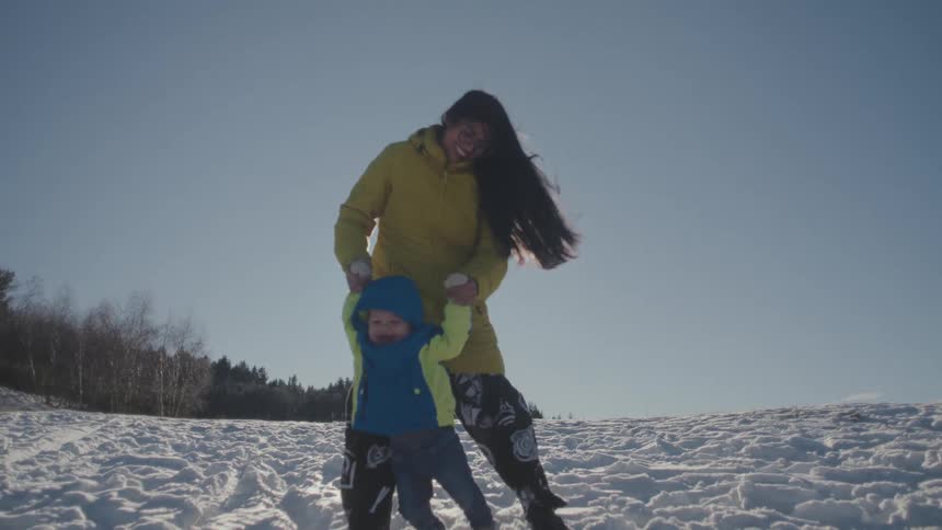 Slow motion shot of a happy mother holding her toddler's hands as he walks and kicks snow on a sunny winter day