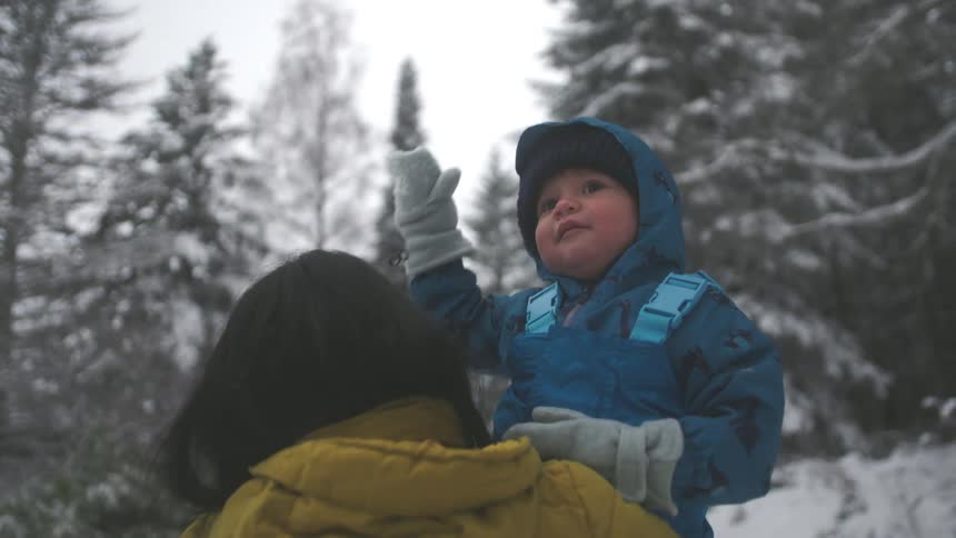 A cute baby bundled in a blue snowsuit sits on their mother's shoulders and looks up in wonder at the falling snow in a winter forest