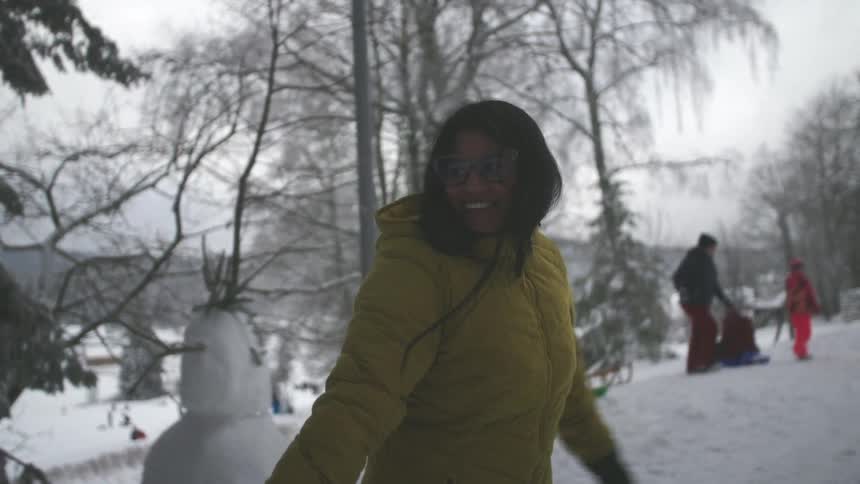 A happy woman in a yellow winter coat smiles while enjoying a snowy day at a park in slow motion