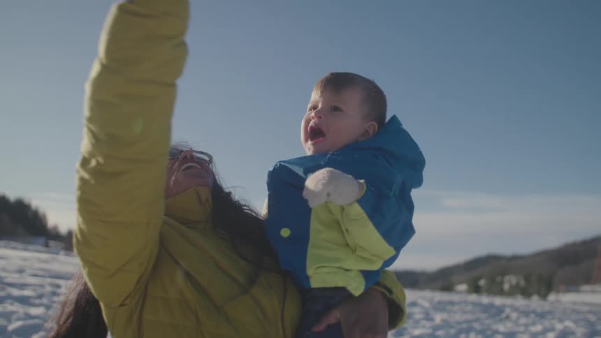A smiling mother holds her baby and plays with the snow, letting it fall from her hand on a bright sunny day in slow motion