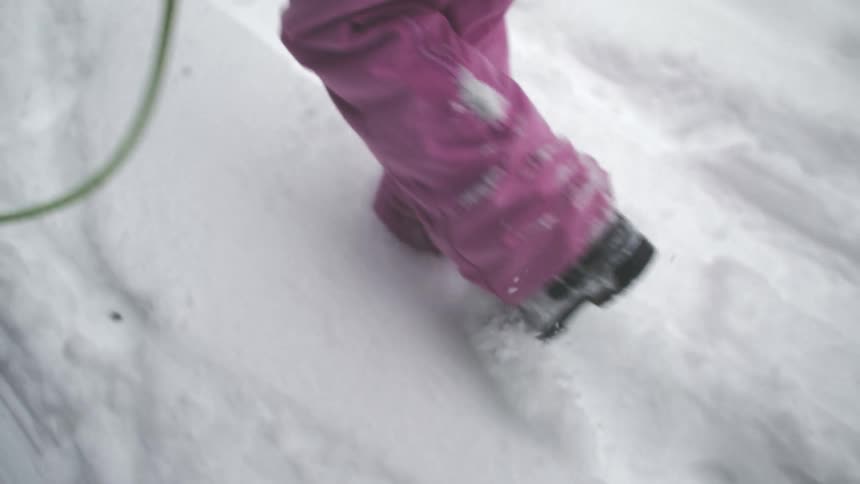 A person in pink snow pants walks uphill through deep snow in slow motion, pulling a sled behind them