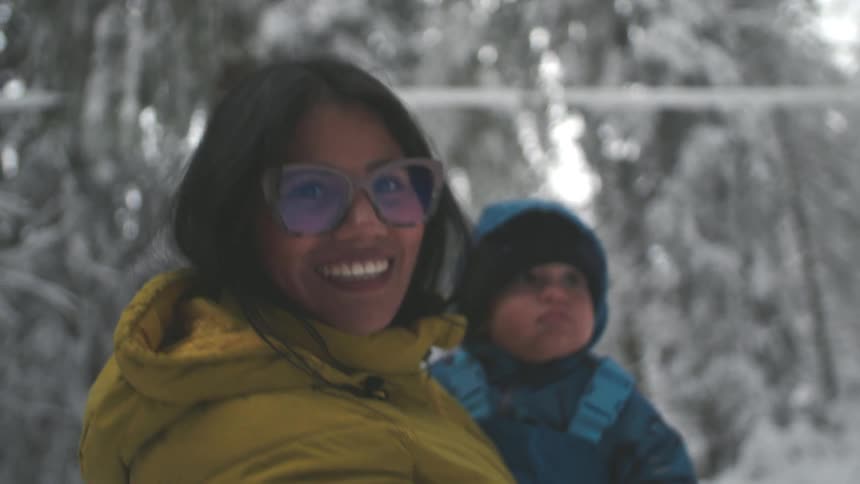A joyful mother smiles while holding her bundled-up toddler in a beautiful snowy winter forest