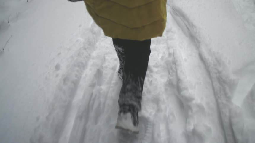 A slow motion, low-angle shot follows a person's legs trudging through deep snow on a winter trail