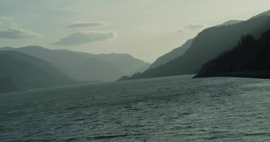 A slow, canted-angle pan across a vast river flowing through a hazy, silhouetted mountain gorge