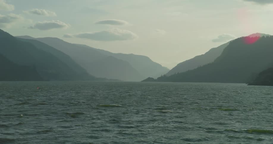 Majestic mountain silhouettes frame a wide, choppy river under a soft, hazy sky