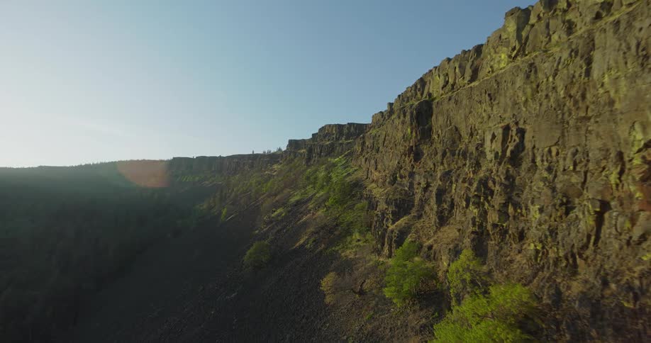 An aerial drone flies smoothly along the edge of a massive, rocky canyon wall under a clear blue sky