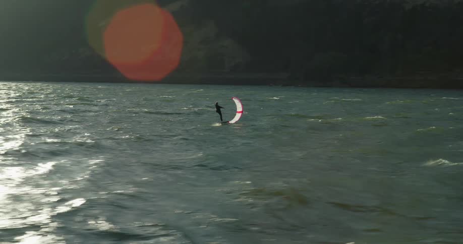 An aerial view follows a lone wing foiler gliding across a shimmering river surrounded by mountains as the sun rises